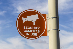 Orange Security Camera In Use Sign on Gray Post and Partly Sunny Background II